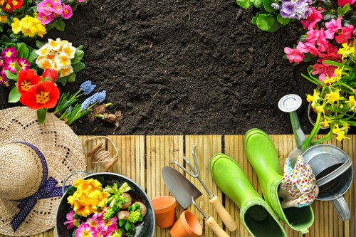The image shows a section of a well-maintained garden with a freshly turned dark soil bed in the upper part of the frame, bordered by a narrow wooden decking area in the lower part. Along the edge of the decking, there are colorful flowering plants including pink, yellow, and purple blooms, creating a vibrant border. On the decking, gardening tools and accessories are arranged, including green rubber rain boots, a metal watering can, a plant spray, small terracotta pots, and a decorative sun hat with a floral ribbon. A small potted plant filled with bright flowers adds to the garden's lush appearance. The environment appears bright, suggesting sunny weather, and the overall scene reflects a neat, organized outdoor space suitable for gardening activities, with elements indicative of attentive lawn and garden care typically offered by services like Lawn Mowing Colindale in the local area around Colindale, London.